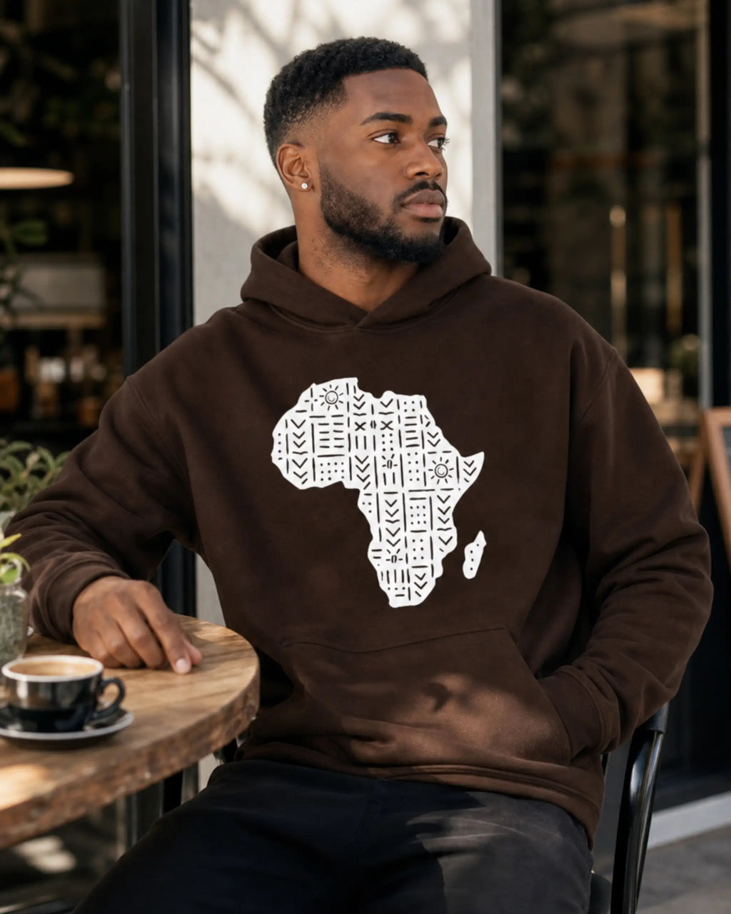 A man wearing a brown hoodie with a white graphic of the African continent filled with patterns, sitting at a wooden table in a cafe.