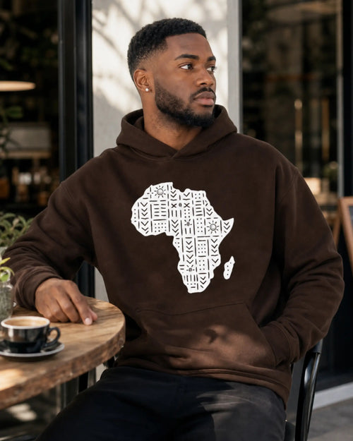 A man wearing a brown hoodie with a white graphic of the African continent filled with patterns, sitting at a wooden table in a cafe.