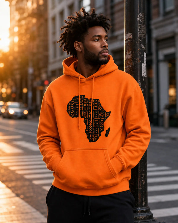 A man wearing a bright orange hoodie with a black Africa map graphic on the front, standing on a city street at sunset.