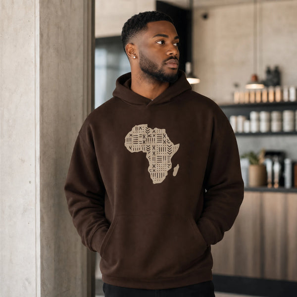 A man wearing a dark brown hoodie with a beige graphic of the African continent made of geometric patterns, standing in a modern indoor setting.